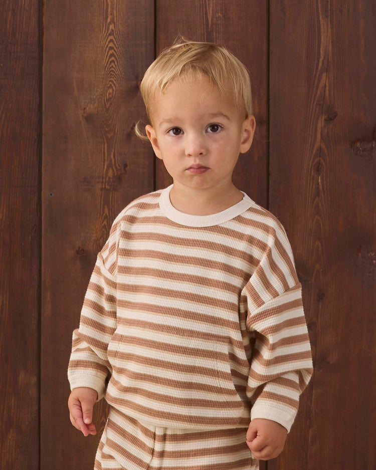 A young child with short blonde hair stands in front of a wooden wall, looking directly at the camera with a neutral expression while wearing the Quincy Mae waffle slouch set in spice stripe.