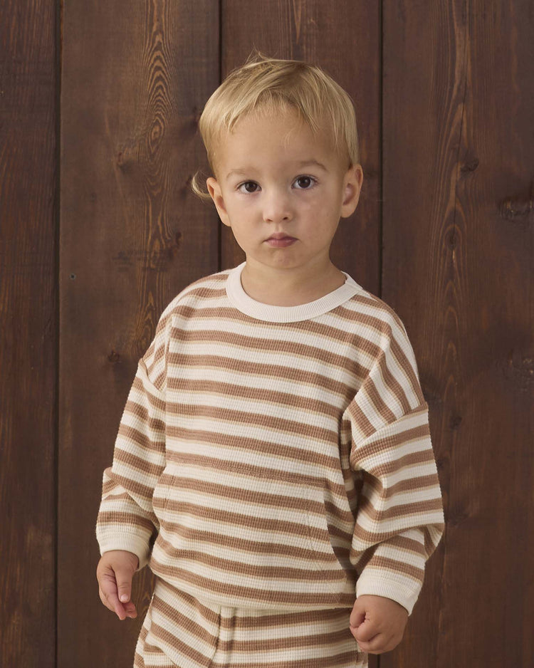 A young child with short blonde hair stands in front of a wooden wall, looking directly at the camera with a neutral expression while wearing the Quincy Mae waffle slouch set in spice stripe.