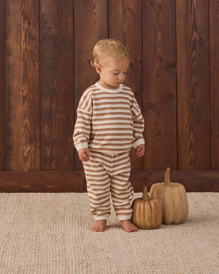 A young child in the quincy mae waffle slouch set in spice stripe, made from organic cotton, stands barefoot on a textured rug beside two decorative pumpkins, with a wooden plank wall in the background.