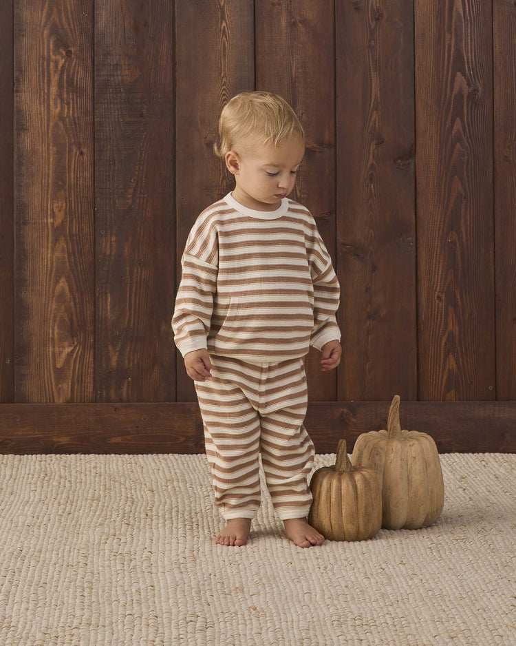 A young child in the quincy mae waffle slouch set in spice stripe, made from organic cotton, stands barefoot on a textured rug beside two decorative pumpkins, with a wooden plank wall in the background.