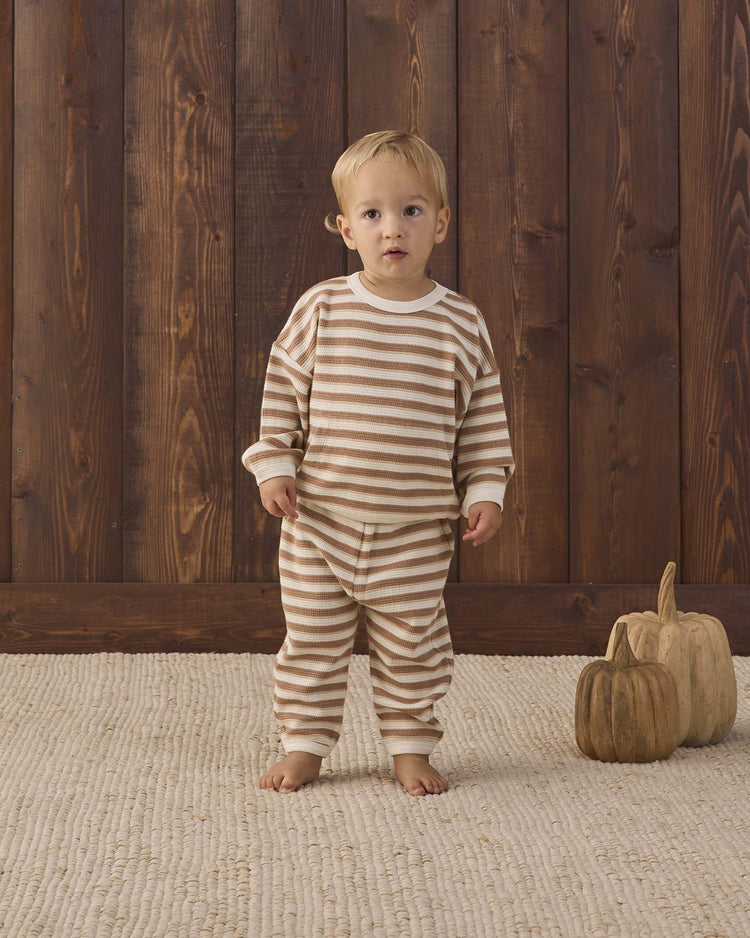 A toddler wearing the quincy mae waffle slouch set in spice stripe, crafted from organic cotton, stands barefoot on a textured rug beside two decorative wooden pumpkins, with a wooden wall as the background.