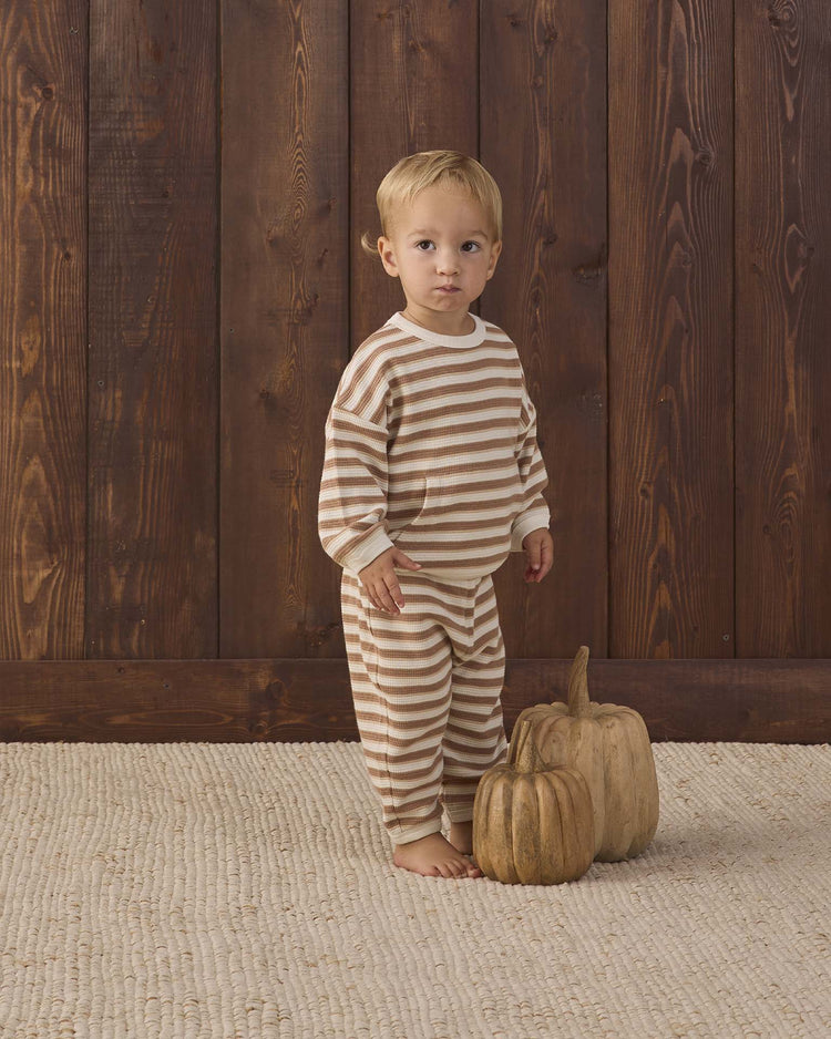 A young child wearing the quincy mae waffle slouch set in spice stripe, crafted from organic cotton, stands barefoot on a textured rug beside two decorative pumpkins, with a wooden wall as the backdrop.