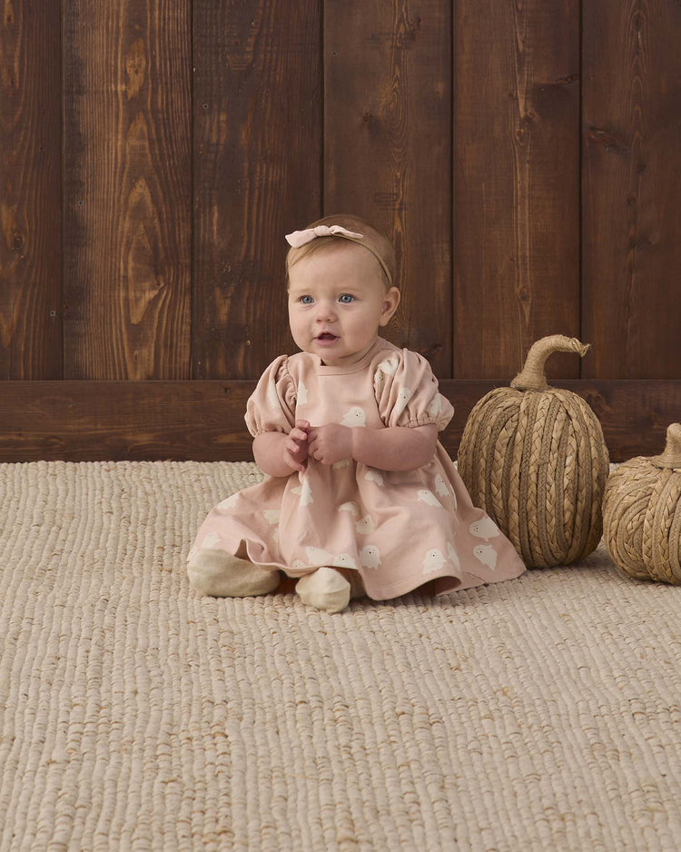 A baby in the quincy mae darla dress in spice stripe and matching headband sits on a textured rug next to two decorative, twine-wrapped pumpkins, with a wooden wall in the background.
