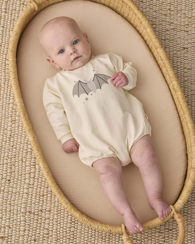 A baby in the quincy mae crewneck bubble romper in baby bat—an organic cotton, cream-colored outfit featuring a playful bat graphic—lies in a woven Moses basket atop a textured beige mat, gazing upward with wide blue eyes.