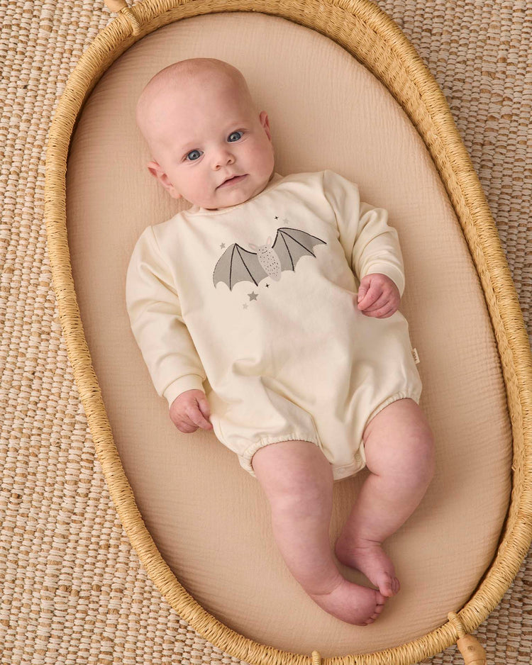 A baby wearing the quincy mae crewneck bubble romper in baby bat lies in a woven basket on a textured beige mat, gazing upward with a calm expression.