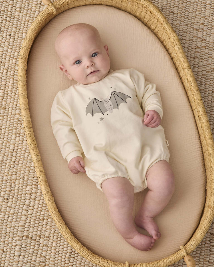 A baby wearing the quincy mae crewneck bubble romper in baby bat lies in a woven basket on a textured beige mat, gazing upward with a calm expression.