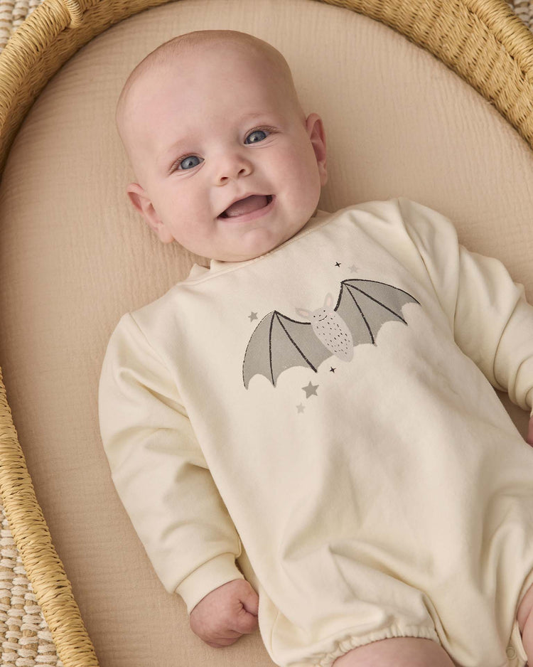 A smiling baby lies in a woven basket, wearing the quincy mae crewneck bubble romper in baby bat—an organic cotton, cream-colored outfit featuring an adorable bat and stars design on the chest.