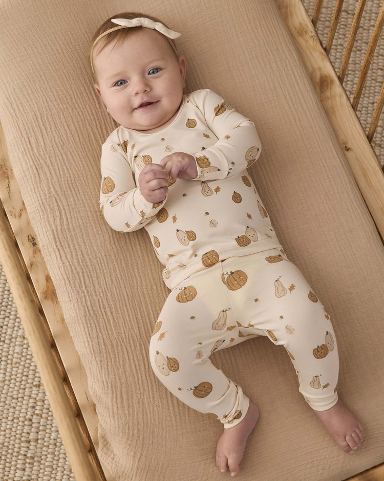 A baby smiles and looks up while lying on a beige blanket in a crib, dressed in the Quincy Mae bamboo pajama set in harvest friends with a matching bow headband.