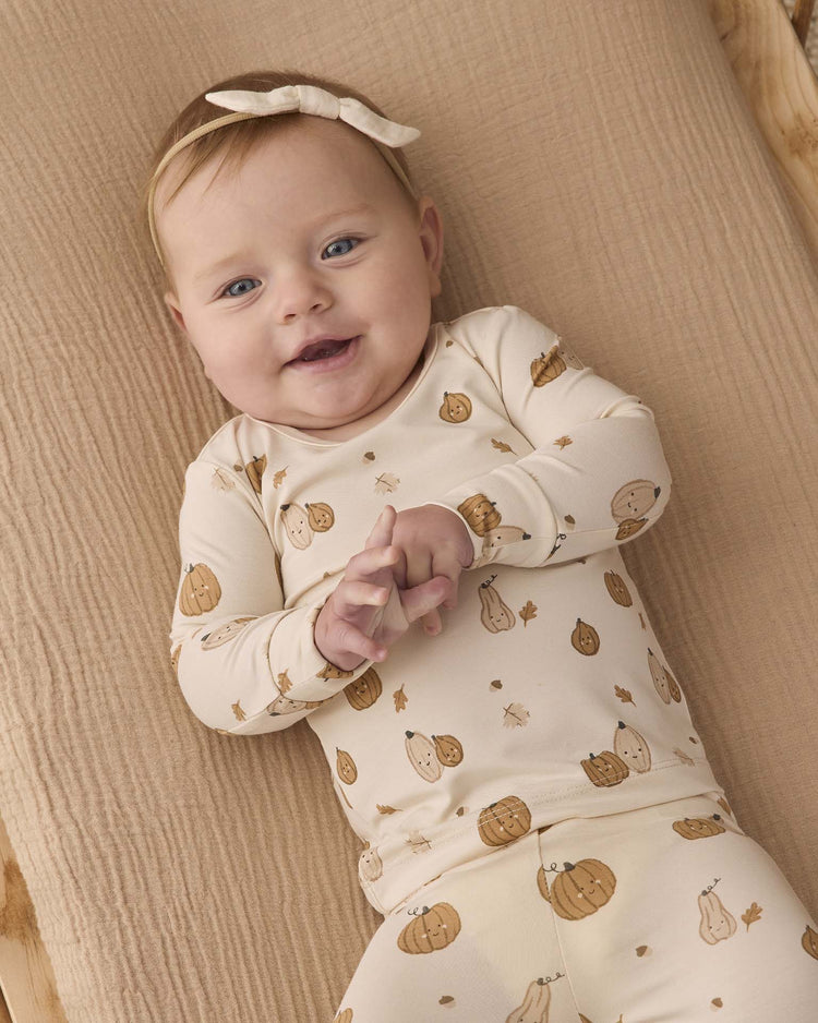 A baby with a light bow headband smiles while lying on a beige blanket, wearing the quincy mae bamboo pajama set in harvest friends.