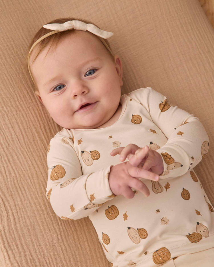 A baby with light hair and blue eyes, wearing the quincy mae bamboo pajama set in harvest friends with a matching headband, lies on a beige textured blanket, smiling softly with hands clasped.