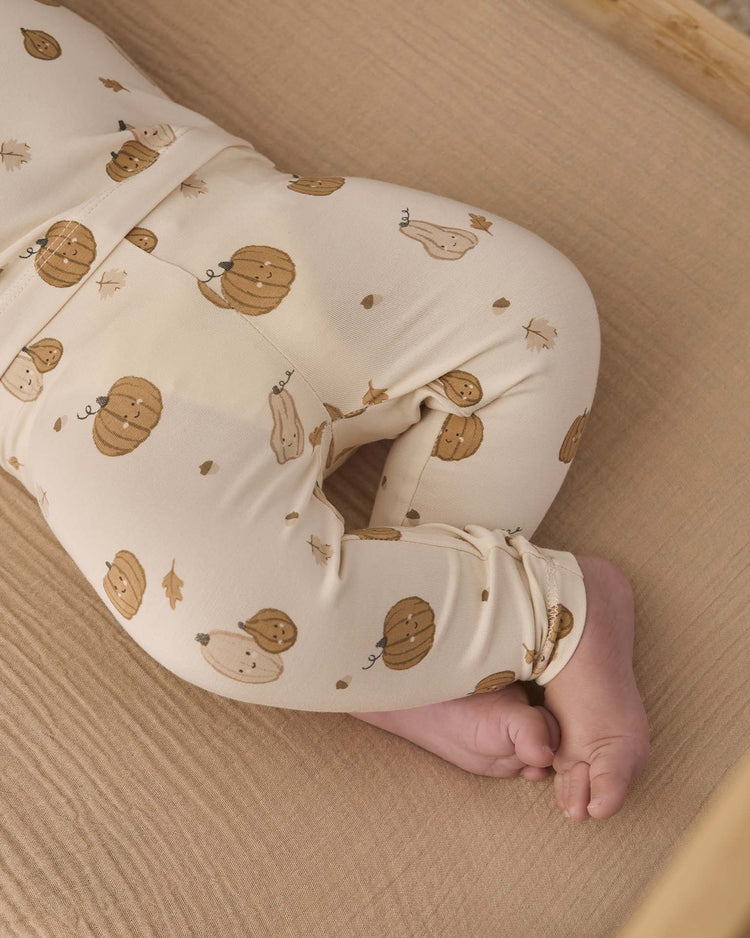 A baby lies on a beige sheet, wearing the Quincy Mae bamboo pajama set in Harvest Friends—a cream-colored slim-fit style featuring brown pumpkin and leaf patterns. Only the lower half of the baby’s body and feet are visible.