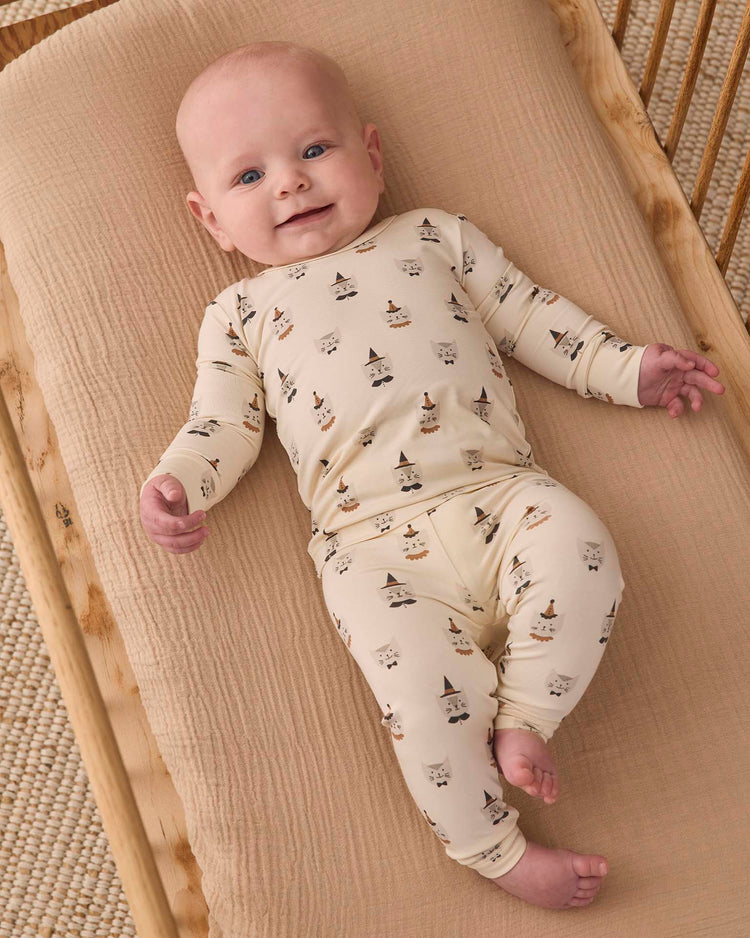 A smiling baby lies on a beige mattress, wearing the quincy mae bamboo pajama set in costume kittens—a slim-fit, cream-colored outfit adorned with playful animal faces and hats—while looking up with one arm bent and legs relaxed.