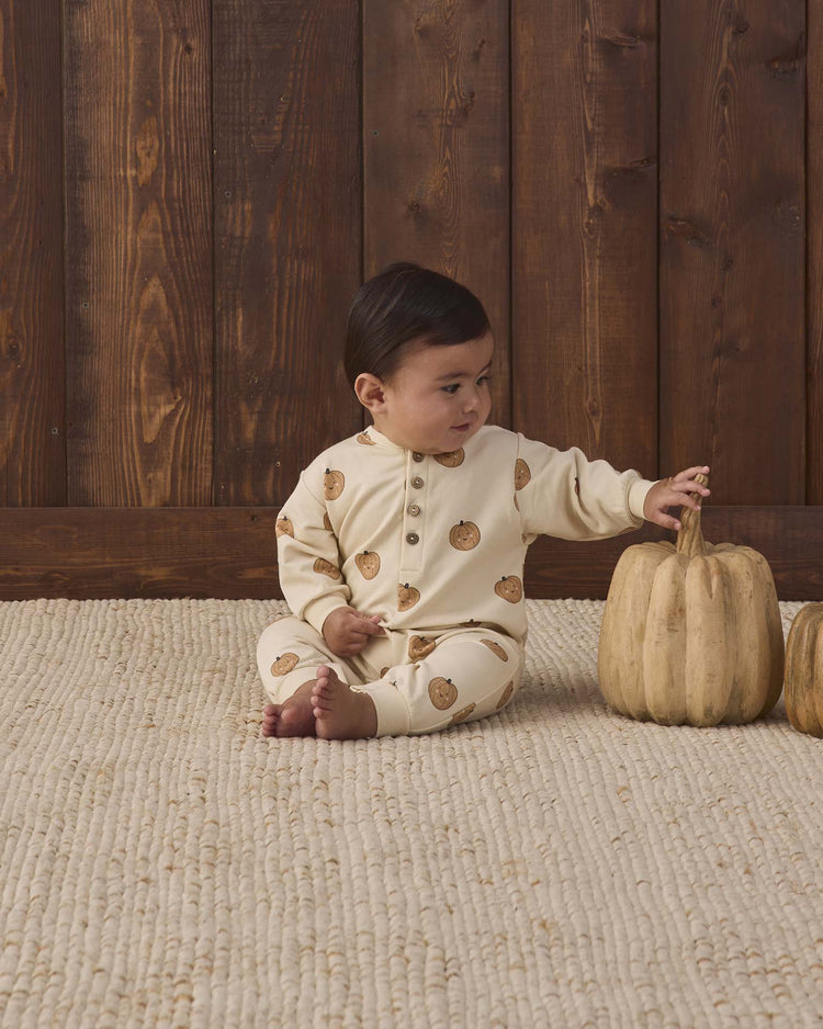 A baby in the quincy mae relaxed fleece jumpsuit in pumpkins, made from cozy organic cotton, sits on a textured rug and reaches for a light-colored pumpkin, with a wooden wall behind them.