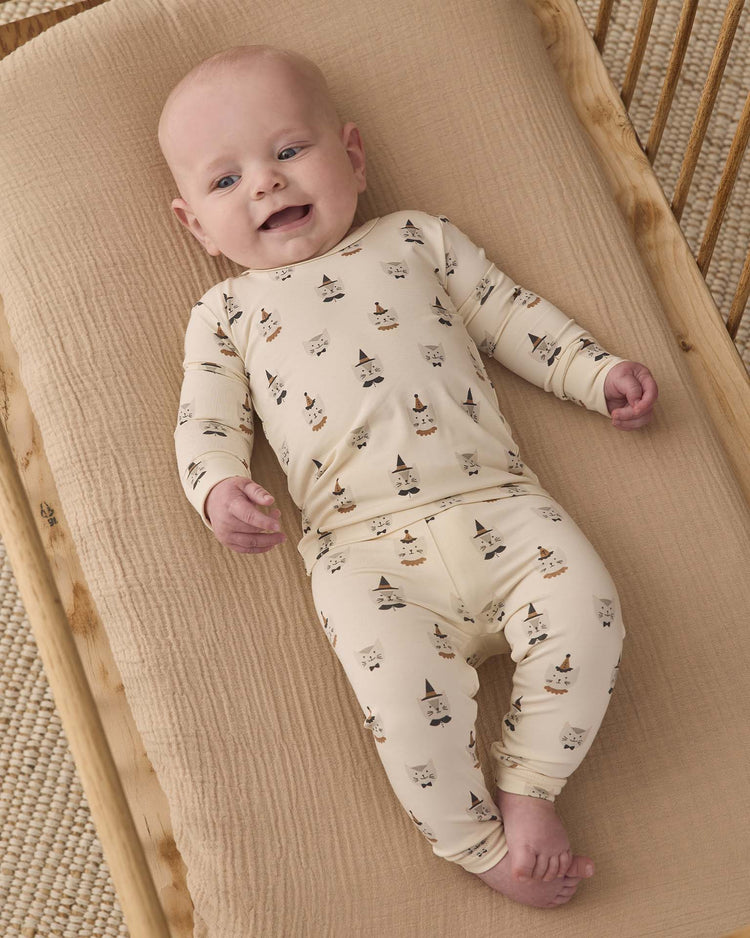 A baby in a Quincy Mae bamboo pajama set in Costume Kittens, featuring a slim fit and long sleeves, lies smiling on a beige padded mat on a wooden crib.