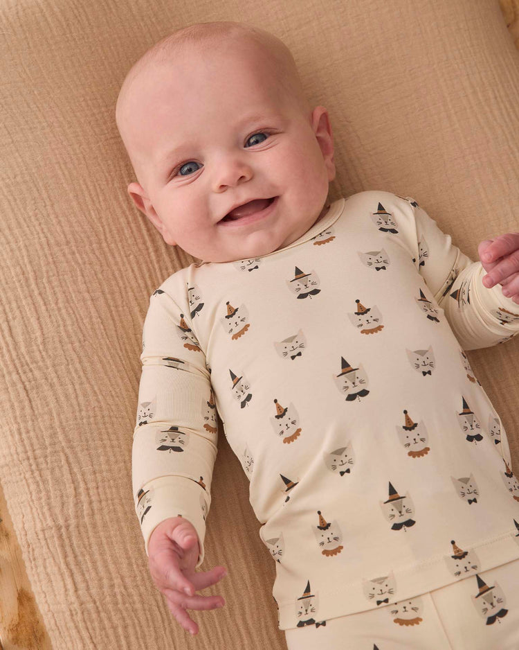 A smiling baby lies on a beige textured blanket, wearing the quincy mae bamboo pajama set in costume kittens, featuring a cream-colored design with cartoon animal faces and hats.