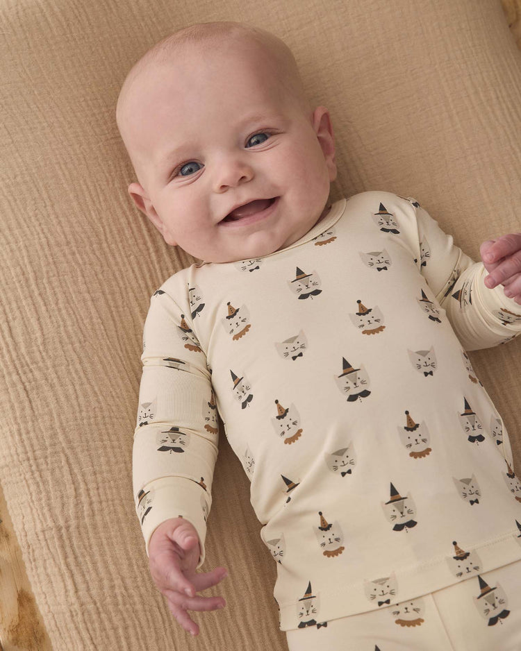 A smiling baby lies on a beige textured blanket, wearing the quincy mae bamboo pajama set in costume kittens, featuring a cream-colored design with cartoon animal faces and hats.