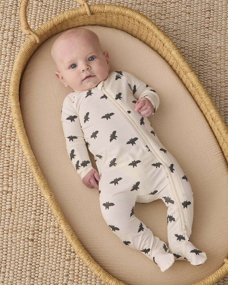 A baby in the quincy mae bamboo zip footie in bats lies in a woven bassinet on a textured rug, looking up with wide blue eyes, cozy in their soft outfit adorned with black bat prints.