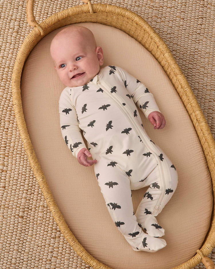 A baby wearing the quincy mae bamboo zip footie in bats lies smiling in a woven bassinet on a textured, woven rug.