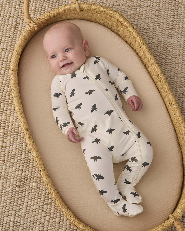 A baby wearing the quincy mae bamboo zip footie in bats lies smiling in a woven bassinet on a textured, woven rug.