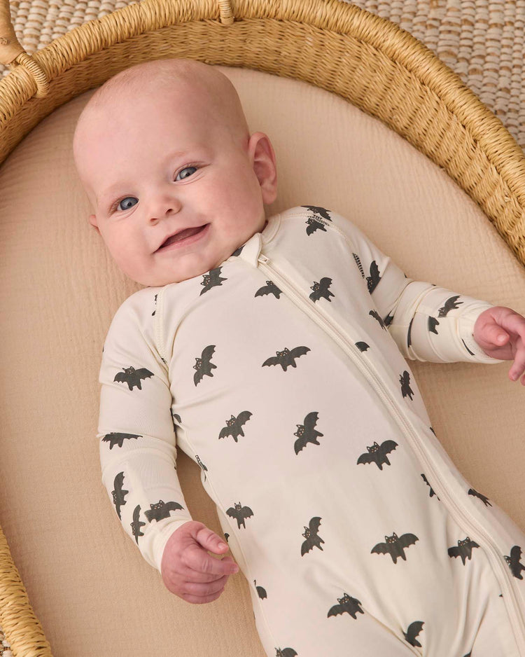 A smiling baby lies in a wicker basket, wearing the quincy mae bamboo zip footie in bats—a cream-colored outfit decorated with black bat prints.