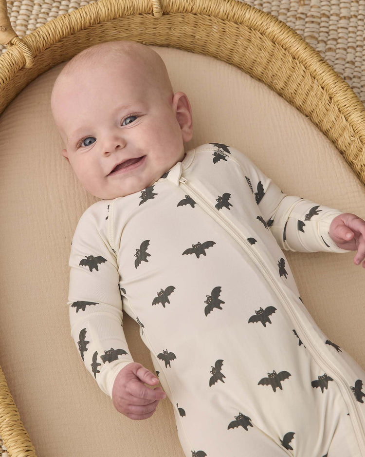 A smiling baby lies in a wicker basket, wearing the quincy mae bamboo zip footie in bats—a cream-colored outfit decorated with black bat prints.
