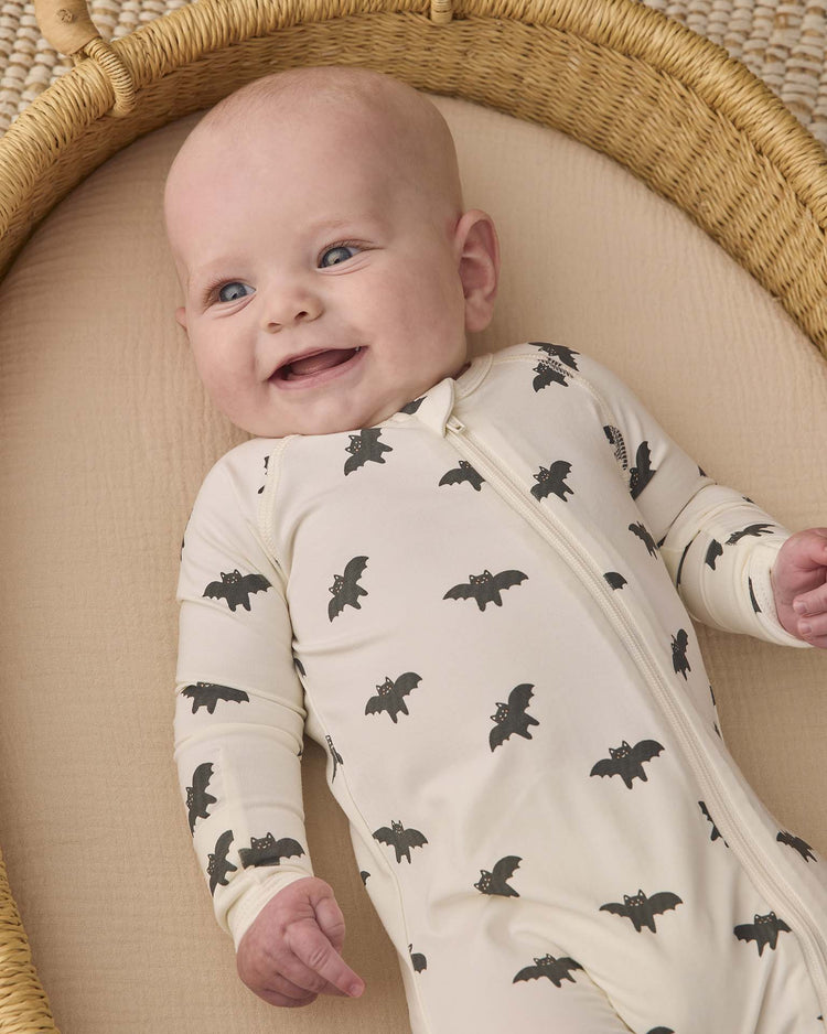 A smiling baby lies in a woven bassinet, wearing the quincy mae bamboo zip footie in bats.