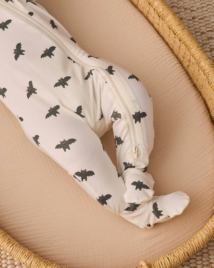 A baby wearing the quincy mae bamboo zip footie in bats, featuring white fabric with black bat prints, lies in a tan woven basket on a beige mattress. Only the lower body and legs of the baby are visible.