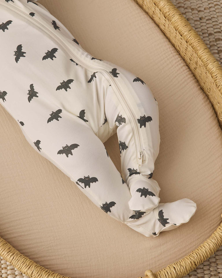 A baby wearing the quincy mae bamboo zip footie in bats, featuring white fabric with black bat prints, lies in a tan woven basket on a beige mattress. Only the lower body and legs of the baby are visible.