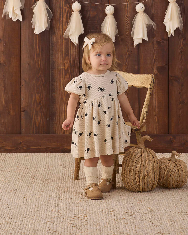 A young child wearing the quincy mae darla dress in bitsy spider—an organic cotton cream dress with black prints—stands by a wooden chair. Two decorative pumpkins and white hanging ornaments are nearby, with a wooden wall and textured rug in the background.