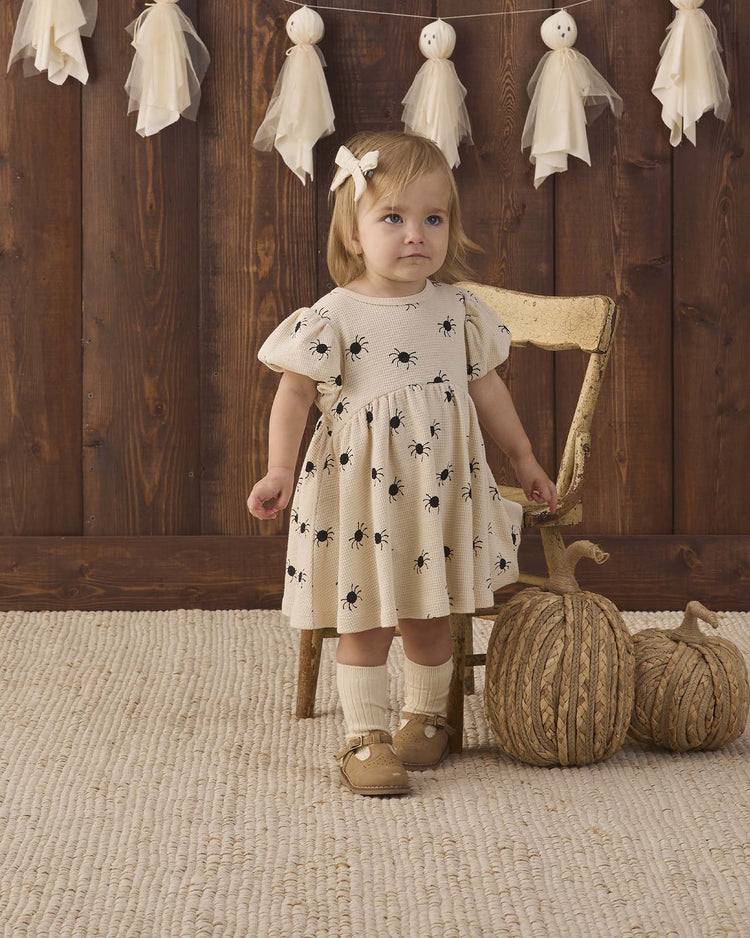 A young child wearing the quincy mae darla dress in bitsy spider—an organic cotton cream dress with black prints—stands by a wooden chair. Two decorative pumpkins and white hanging ornaments are nearby, with a wooden wall and textured rug in the background.