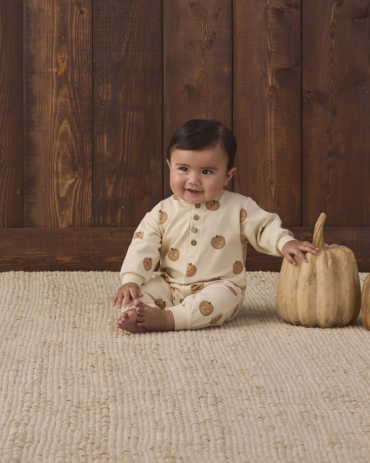A baby wearing the quincy mae relaxed fleece jumpsuit in pumpkins sits on a woven rug, smiling, with one hand resting on a decorative pumpkin. A wooden wall and another pumpkin are visible in the background.