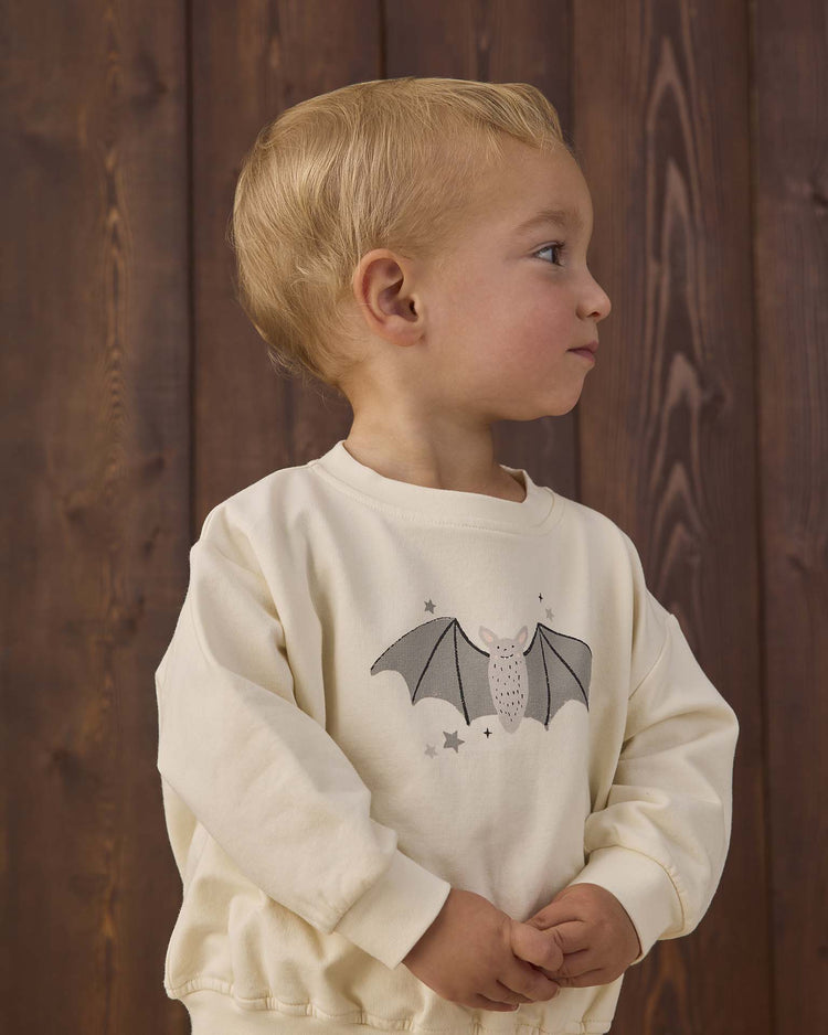 A young child with blond hair stands in front of a wooden wall, wearing the quincy mae relaxed fleece sweatshirt in baby bats—a cream-colored organic cotton top featuring a baby bat and stars graphic on the front. The child is looking to the right with hands clasped together.