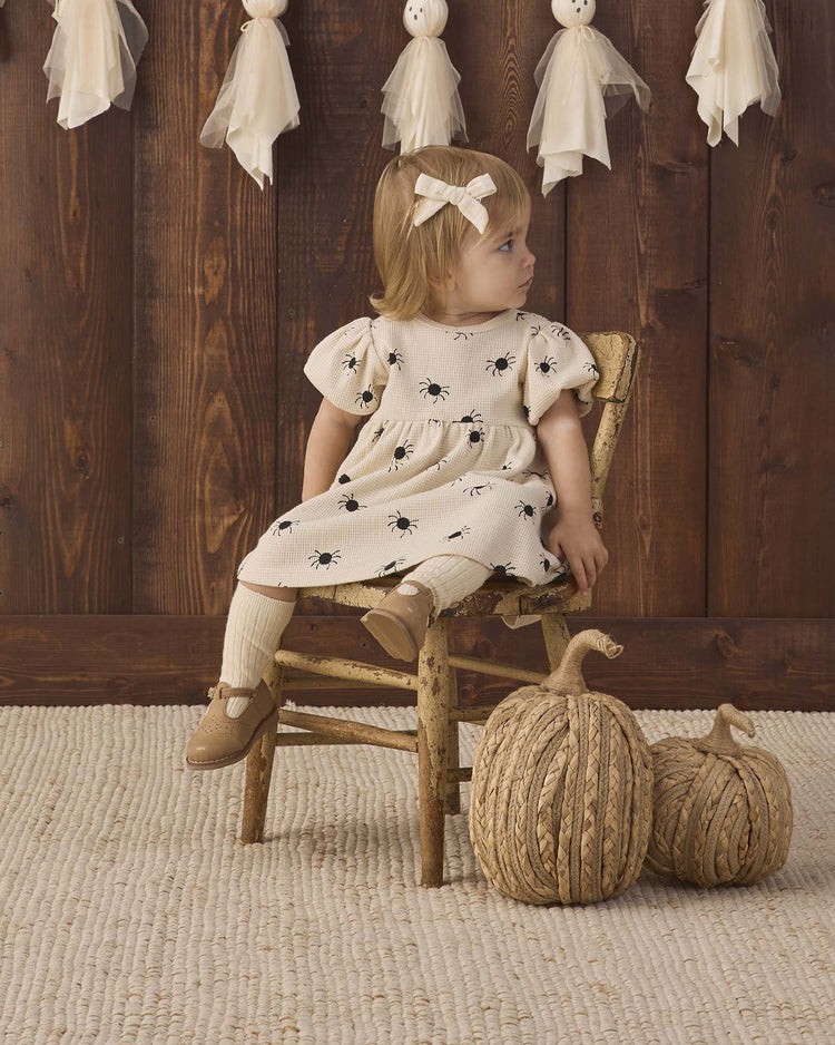 A toddler wearing the quincy mae darla dress in bitsy spider, made from cream organic cotton with black prints, sits on a rustic wooden chair and gazes off to the side. Two woven pumpkins rest on the floor nearby, while ghost decorations hang from a wooden wall in the background.