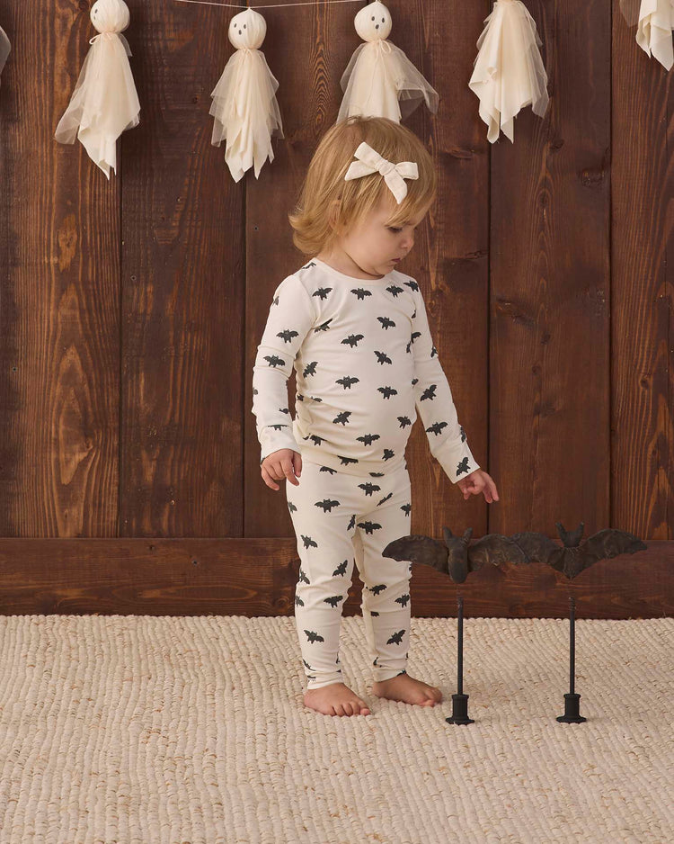 A toddler wears the quincy mae bamboo pajama set in bats, standing barefoot on a rug and looking at two decorative bats on stands, while ghost decorations hang above against a wooden wall.