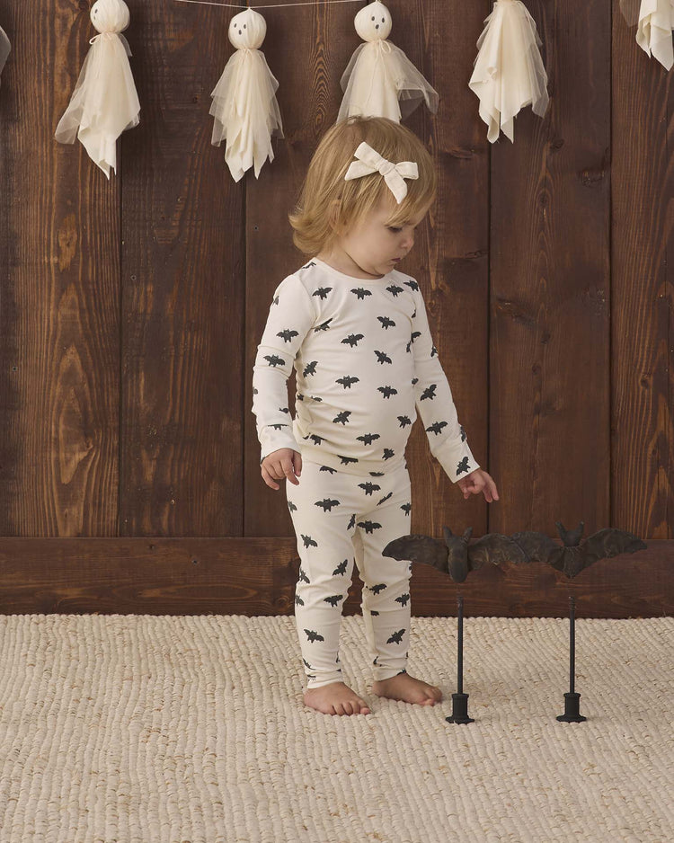 A toddler wears the quincy mae bamboo pajama set in bats, standing barefoot on a rug and looking at two decorative bats on stands, while ghost decorations hang above against a wooden wall.