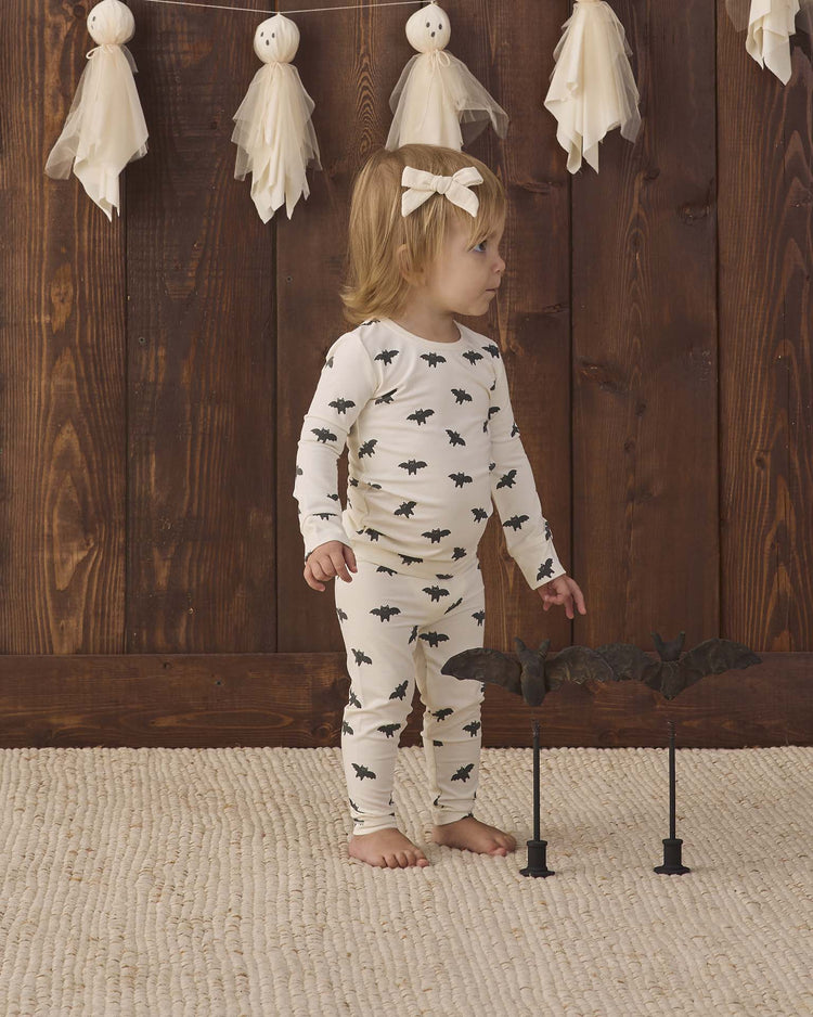 A toddler wearing the quincy mae bamboo pajama set in bats stands on a textured rug, gazing to the side. Behind her, a wooden wall is decorated with ghost ornaments, and two bat props on stands are positioned in front of her.