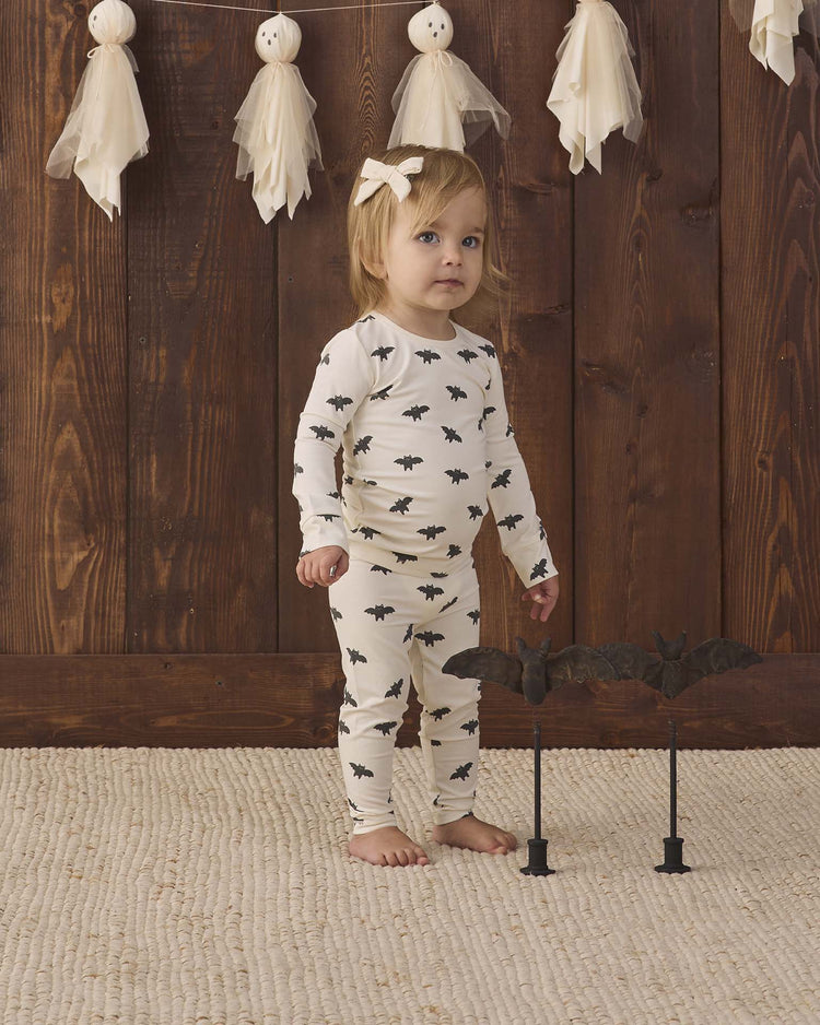 A toddler wearing the quincy mae bamboo pajama set in bats stands on a textured rug with a bat decoration nearby, in front of a wooden wall adorned with white ghost garlands.