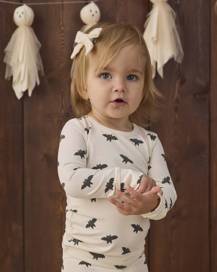 A toddler with light brown hair and a bow wears the quincy mae bamboo pajama set in bats, a slim-fit, cute bat-printed outfit. She stands in front of a wooden wall decorated with ghost-shaped garlands.