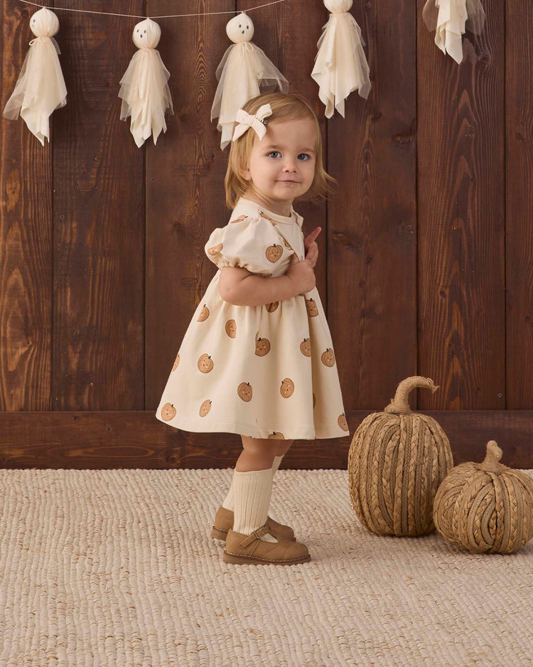 A young child wearing the quincy mae darla dress in pumpkins, made from organic cotton and featuring pumpkin prints, stands on a textured rug. She accessorizes with a hair bow, socks, and brown shoes. Two decorative pumpkins and tassel garlands decorate the background against a wooden wall.