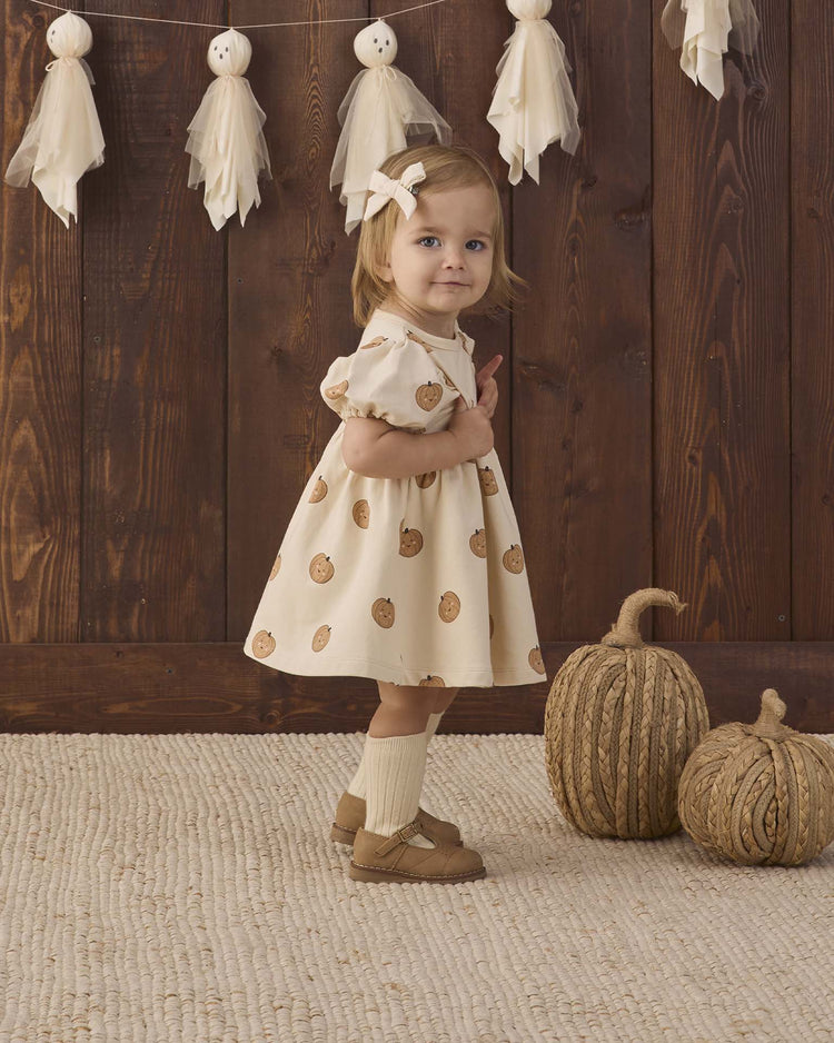 A young child wearing the quincy mae darla dress in pumpkins, made from organic cotton and featuring pumpkin prints, stands on a textured rug. She accessorizes with a hair bow, socks, and brown shoes. Two decorative pumpkins and tassel garlands decorate the background against a wooden wall.