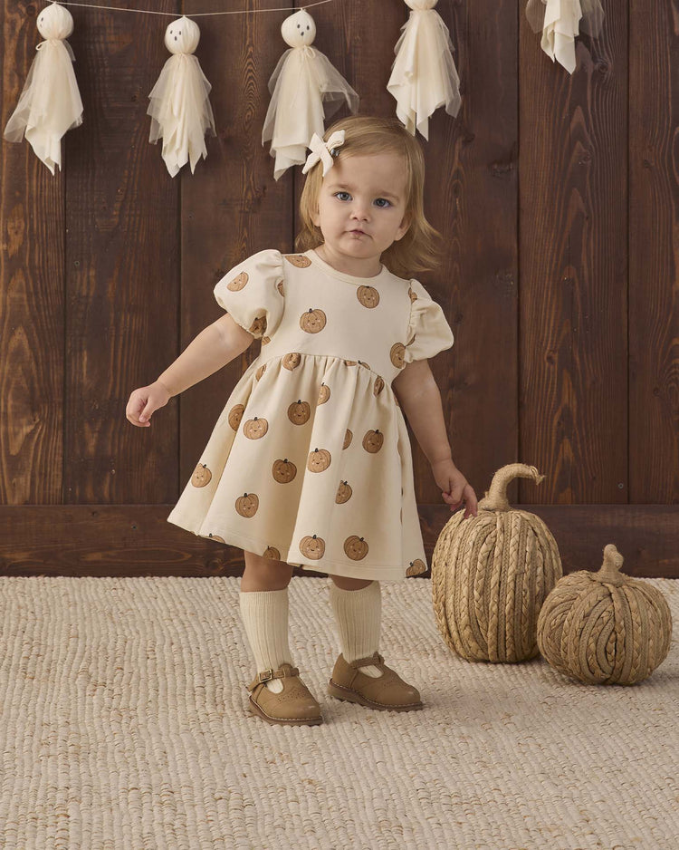 A toddler wearing the quincy mae darla dress in pumpkins—a cream fleece dress with a pumpkin print—stands on a beige rug near two textured pumpkin decorations, with a wooden wall and a hanging tassel garland in the background.
