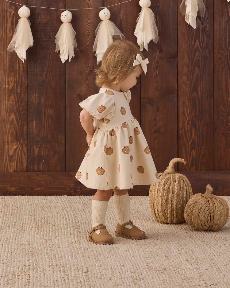 A young child stands indoors near two decorative pumpkins, wearing the quincy mae darla dress in pumpkins—a cream organic cotton baby dress with a pumpkin print—while ghost decorations hang on the wooden wall behind her.