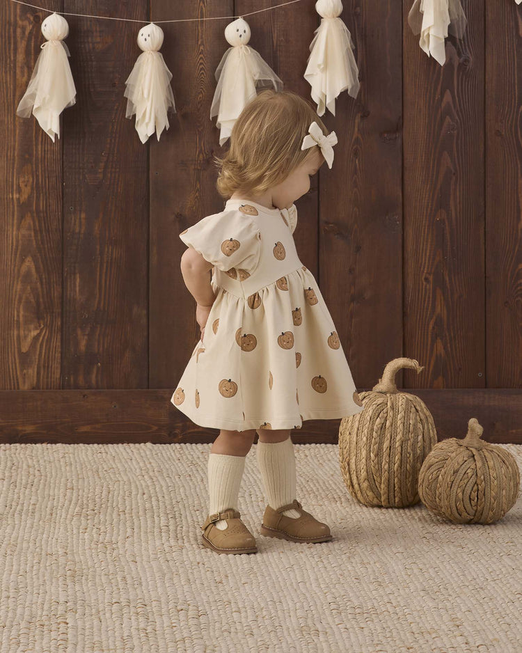 A young child stands indoors near two decorative pumpkins, wearing the quincy mae darla dress in pumpkins—a cream organic cotton baby dress with a pumpkin print—while ghost decorations hang on the wooden wall behind her.