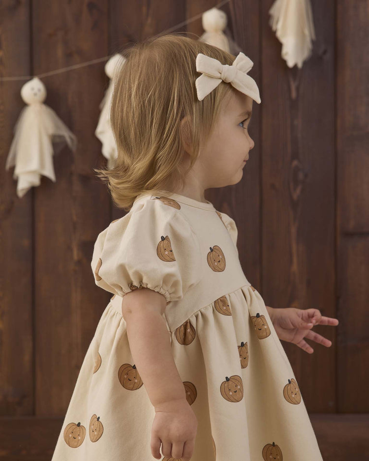 A young child wearing the quincy mae darla dress in pumpkins—a cream fleece dress with a cheerful pumpkin print and a matching bow—stands sideways against a wooden backdrop decorated with hanging ghosts.