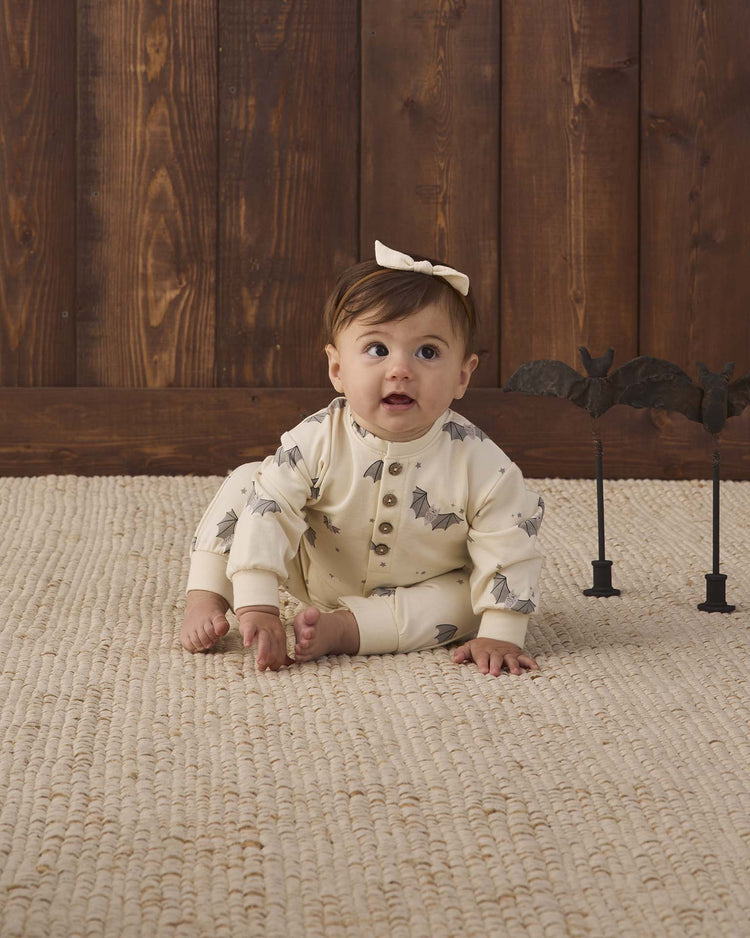 A baby wearing the quincy mae relaxed fleece jumpsuit in baby bats, made from soft organic cotton and featuring a cream bat print, with a white bow, sits on a textured rug in front of a wooden wall while two small black bat decorations rest nearby.