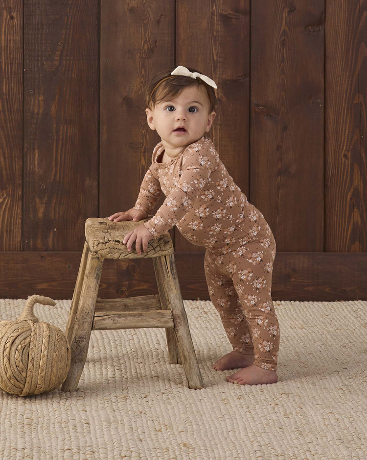 A baby wearing the quincy mae bamboo pajama set in fall floral stands holding a small wooden stool, with a ball of thick rope nearby, on a textured rug and wooden wall background.