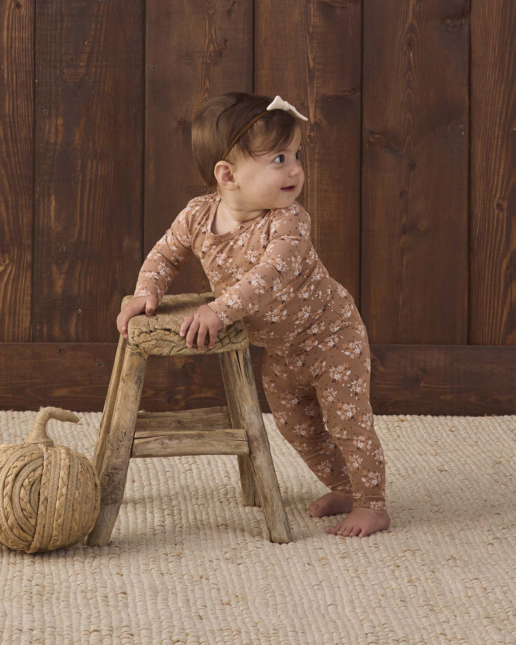 A baby wearing the Quincy Mae bamboo pajama set in fall floral stands holding a small wooden stool, looking to the side. The background features a wooden wall and a cream woven rug with a large ball of rope on the floor.