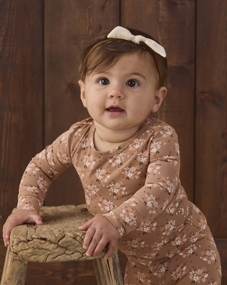 A baby wearing the quincy mae bamboo pajama set in fall floral stands holding onto a rustic wooden stool, with a dark wooden background behind, and is accessorized with a cream bow headband.
