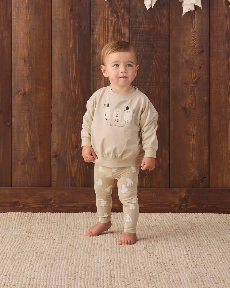 A toddler stands barefoot on a beige rug, dressed in light-colored animal print pajamas and the quincy mae relaxed fleece sweatshirt in trick or treat, in front of a wooden wall.