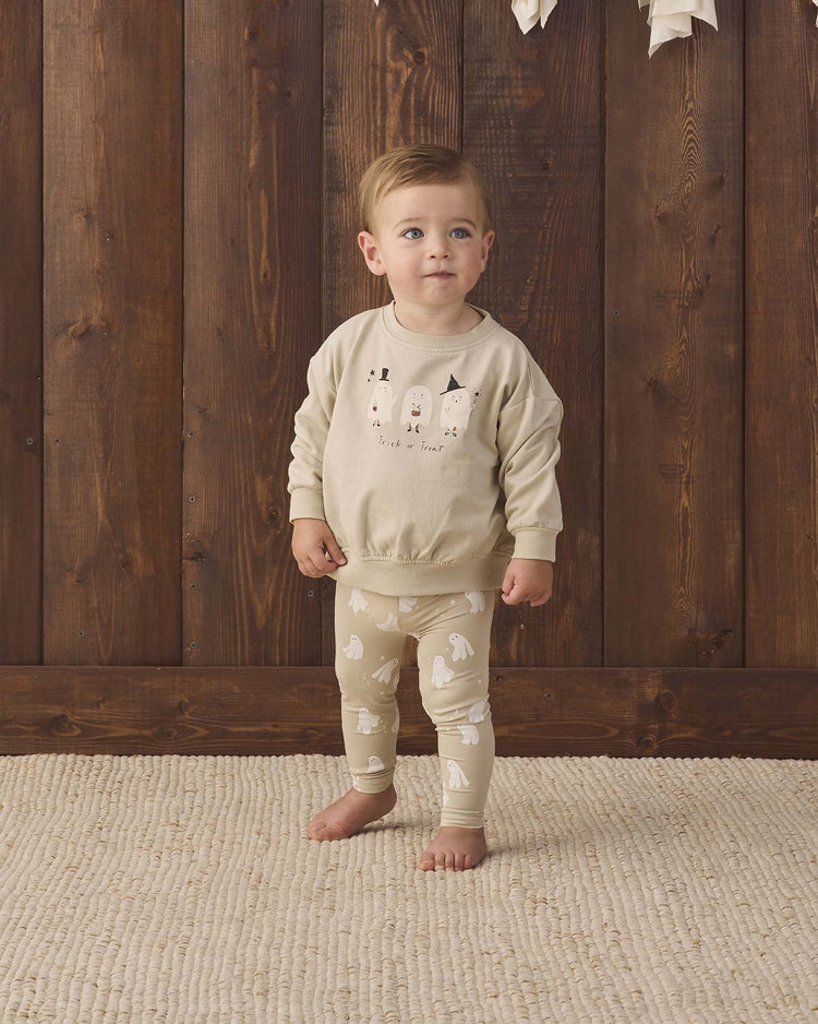 A toddler stands barefoot on a beige rug, dressed in light-colored animal print pajamas and the quincy mae relaxed fleece sweatshirt in trick or treat, in front of a wooden wall.